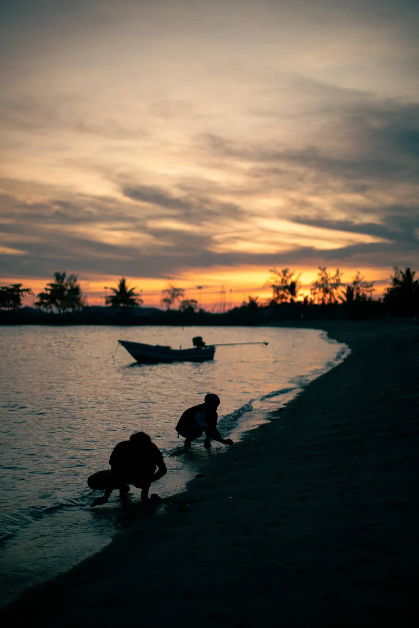 Sunset over Mae Haad beach, Ko Pha Ngan — a longtail boat on still water, palms in silhouette against an orange sky, a figure crouched at the waterline.