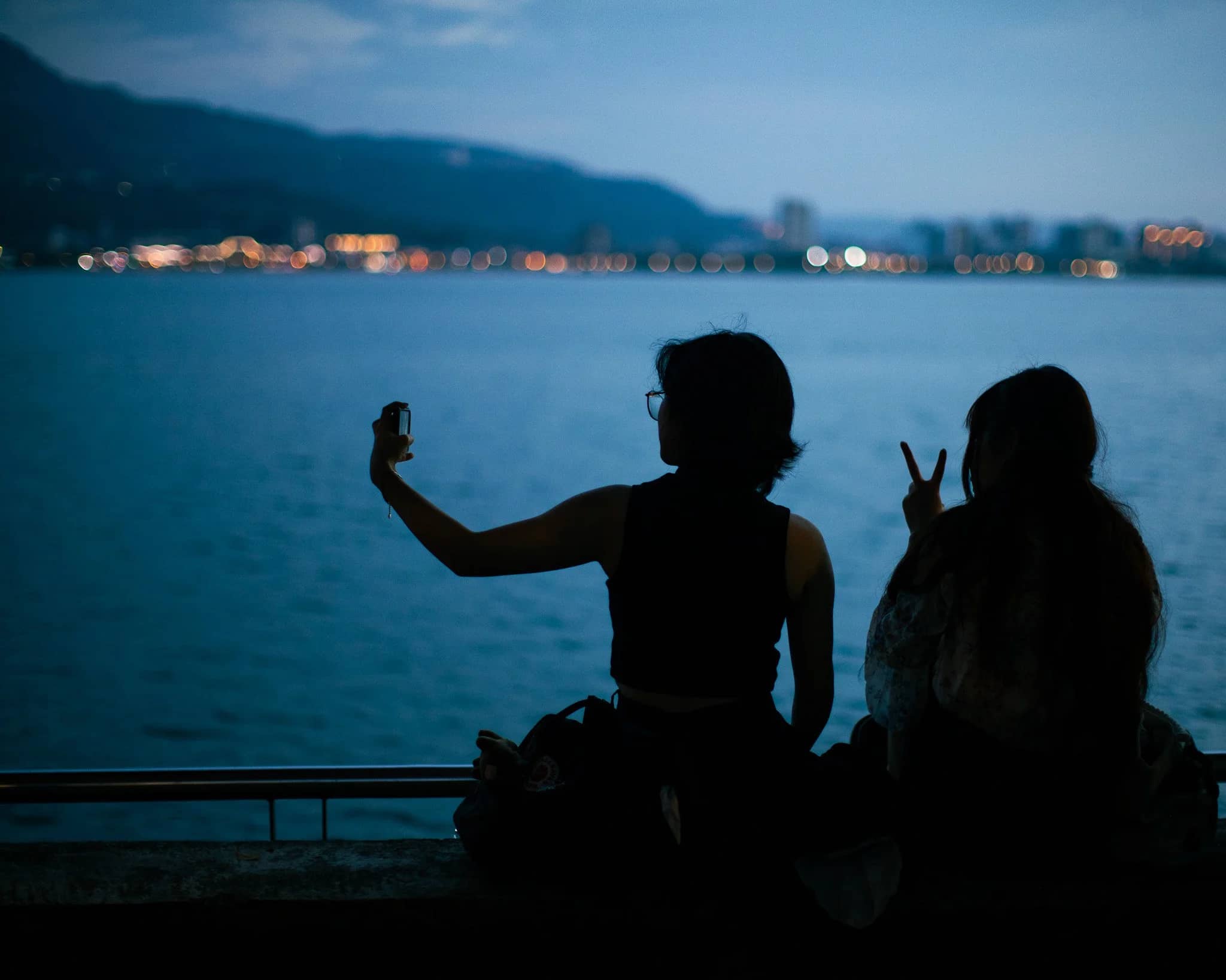 Two figures in silhouette on a Taiwanese waterfront at blue hour, one holding up a phone, city lights strung along the far shore.