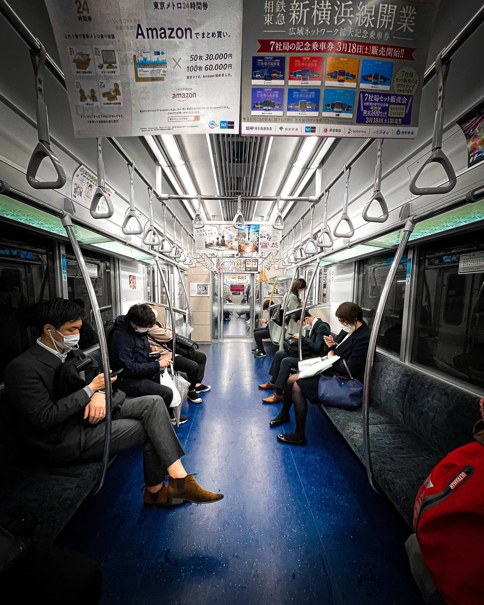 The interior of a Tokyo subway carriage — commuters seated along both sides under fluorescent light, hand straps in a row down the centre.
